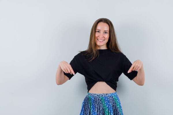 Young lady pointing down in blouse, skirt and looking joyful. front view.