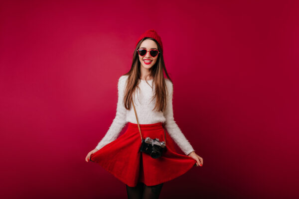 Enthusiastic long haired female photographer playing with red skirt. portrait of glad young woman in trendy hat posing with camera..